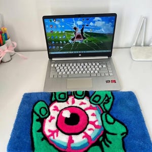 May include: A blue rug with a green hand holding a pink and white eyeball design. The rug is placed on a white desk with a laptop displaying a cracked screen. A white router is also on the desk.