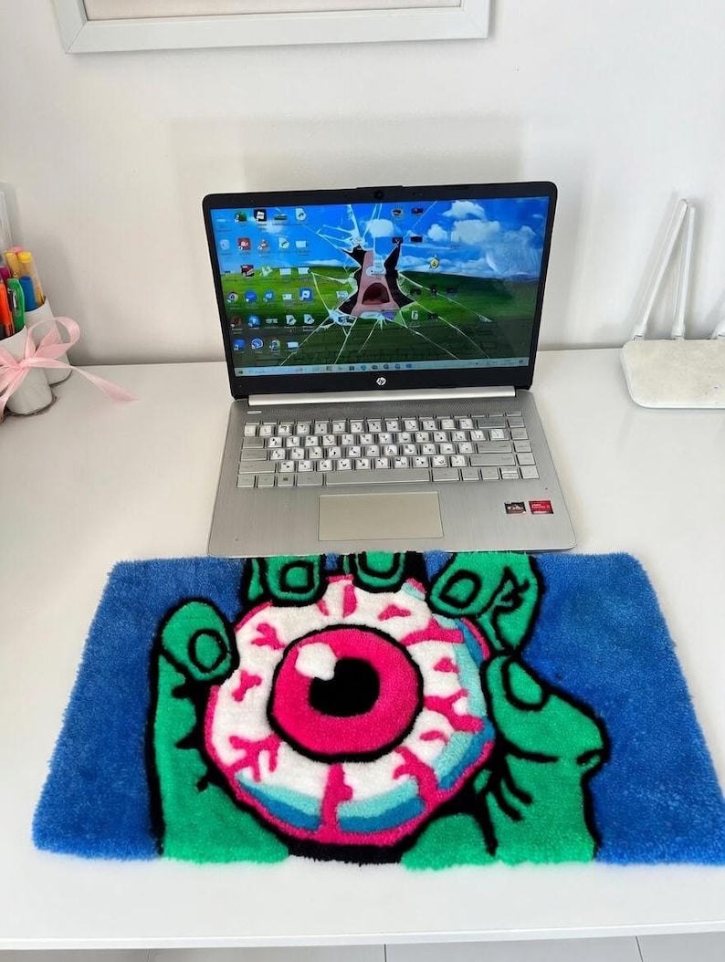 May include: A blue rug with a green hand holding a pink and white eyeball design. The rug is placed on a white desk with a laptop displaying a cracked screen. A white router is also on the desk.