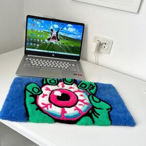 May include: A blue rug with a cartoon eye design. The eye is pink, white, and black, held by green hands. The rug is rectangular and sits on a white surface. A laptop with a cracked screen is in the background.