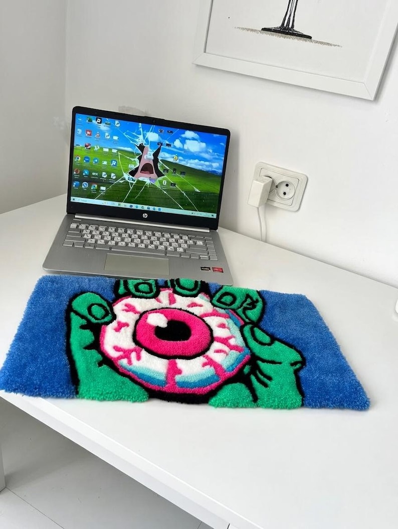 May include: A blue rug with a cartoon eye design. The eye is pink, white, and black, held by green hands. The rug is rectangular and sits on a white surface. A laptop with a cracked screen is in the background.
