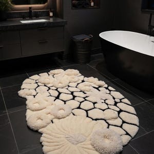 May include: A cream-colored, textured rug with a unique design. The rug features a pattern of interconnected black shapes and raised, circular cream-colored elements. The rug is placed on a dark tiled floor in a bathroom setting.