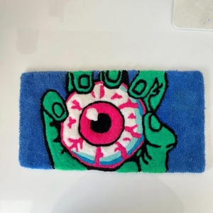 May include: A rectangular rug featuring a green hand holding a large eyeball. The eyeball has a pink iris, a black pupil, and white and blue details. The background is blue, and the rug is made of a soft, textured material.