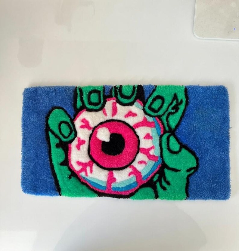 May include: A rectangular rug featuring a green hand holding a large eyeball. The eyeball has a pink iris, a black pupil, and white and blue details. The background is blue, and the rug is made of a soft, textured material.