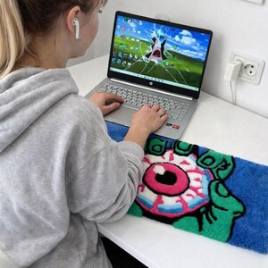 May include: A blue rectangular rug with a cartoon eye design. The eye has a pink iris, white sclera, and green and blue accents. A laptop with a cracked screen is in the background.