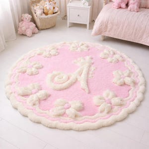 May include: A round, light pink rug featuring a large, raised, cream-colored letter A in the center. The rug is decorated with a repeating pattern of raised, cream-colored bows around the perimeter and has a scalloped, cream-colored edge.
