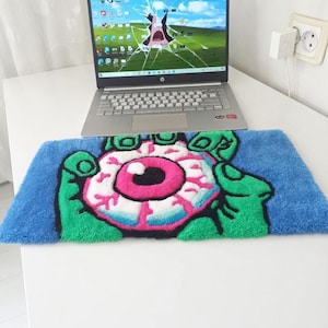 May include: A blue, green, pink, and white rug featuring a cartoon eyeball design held by green hands. The rug is rectangular and placed in front of a laptop with a cracked screen. The laptop is silver and sits on a white surface.