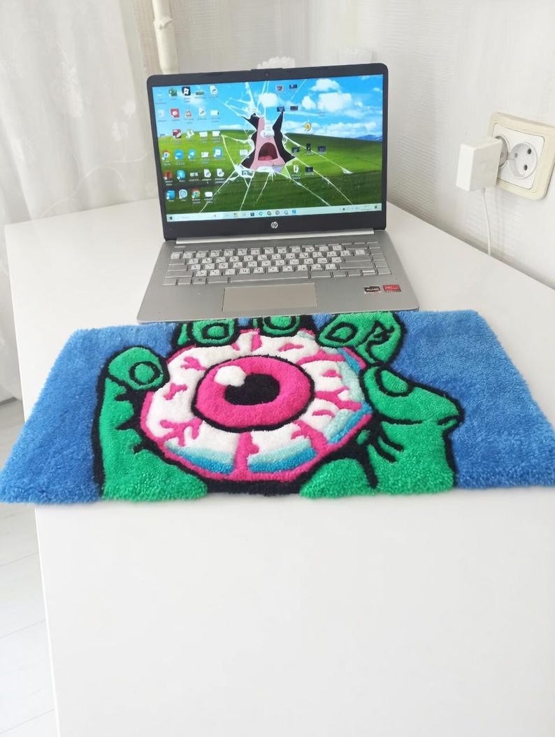 May include: A blue, green, pink, and white rug featuring a cartoon eyeball design held by green hands. The rug is rectangular and placed in front of a laptop with a cracked screen. The laptop is silver and sits on a white surface.