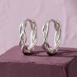 May include: A pair of silver hoop earrings featuring a polished, braided metal design. The circular hoops have a continuous twisted texture that creates a woven appearance, with a hinged closure mechanism visible at the top of each earring.