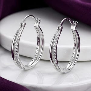 May include: A pair of silver-toned hoop earrings featuring a double-band design. Each earring is adorned with a row of small, clear, sparkling stones embedded along the front-facing band, secured with a hinged post closure at the top.