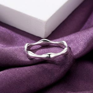 May include: A silver ring with a flowing, wavy design. The ring is made of a shiny metal and rests on a purple satin fabric. The ring's design is organic, with a smooth, polished finish. The ring is a piece of jewellery.