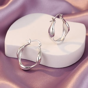 Puede incluir: Un par de pendientes de aro plateados con un diseño ondulado. Los pendientes se exhiben sobre una superficie rectangular blanca sobre un fondo de tela de satén púrpura. Los pendientes tienen una superficie pulida y reflectante.