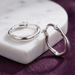 May include: A pair of silver hoop earrings with an irregular, organic, wavy shape. The metal surface is smooth and polished, reflecting light. Each earring features a hinged closure mechanism and is stamped with the S925 hallmark for sterling silver.