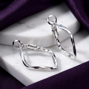 May include: A pair of silver hoop earrings featuring a twisted, diamond-shaped design. Each earring has a hinged post closure mechanism. The metal has a polished, reflective finish, and the earrings are positioned to show their unique geometric shape.