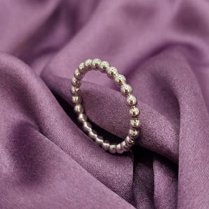 May include: A silver ring with a beaded design. The ring is composed of small, evenly spaced spheres, creating a textured surface. The ring is displayed on a purple satin fabric.