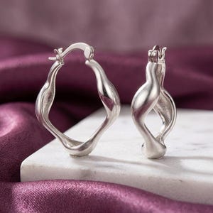 May include: A pair of silver hoop earrings with a distinct, irregular, wavy, and organic shape. The earrings feature a polished metallic finish and a hinged closure mechanism at the top, showcasing a sculptural design with smooth, flowing curves.