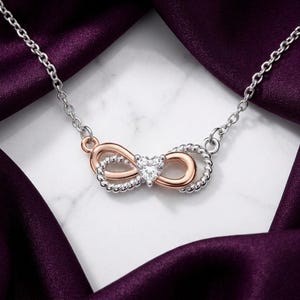 Puede incluir: Un collar con cadena plateada y un colgante central en forma de infinito. El colgante combina metal liso en tono oro rosa con metal plateado trenzado, con una pequeña piedra preciosa transparente en forma de corazón en el centro.