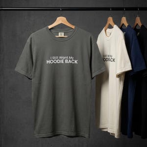 Puede incluir: Varias camisetas cuelgan de una percha. La primera es una camiseta gris oscuro con el texto "I Still Want My HOODIE BACK" en blanco. Otras camisetas son crema, azul marino y negro. Todas las camisetas tienen cuello redondo y mangas cortas.