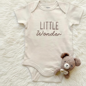 May include: A cream-colored baby onesie with the words "LITTLE Wonder" in brown script. A small, crocheted brown teddy bear is next to the onesie. The onesie has short sleeves and snaps at the bottom.