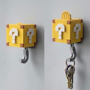 May include: A yellow Lego-style question mark block key holder with a coin on top. The block has white question marks on the front and sides. A silver hook extends from the bottom, holding a set of keys. The key holder is mounted on a white wall.