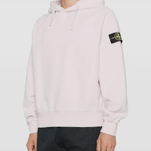 以下が含まれることがあります： ドローストリングフードと長袖が付いた、薄いピンク色のパーカー。左袖には小さな黒と黄色のパッチが付いています。柔らかい素材で作られており、ゆったりとしたフィット感です。