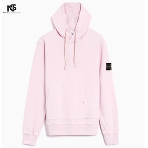 Peut inclure: Un sweat à capuche rose clair avec une capuche à cordon et une poche kangourou. Le sweat à capuche a des manches longues et un petit patch noir et vert sur la manche gauche. Le logo de la marque "NOYAS STITCH STUDIO" est dans le coin supérieur gauche.