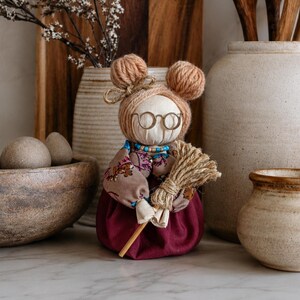 Puede incluir: Una muñeca de tela con cabello de lana en dos moños, gafas de montura metálica, una blusa de manga larga con estampado floral y una falda roja oscura lisa. La muñeca sostiene un pequeño manojo de paja seca y lleva un collar de cuentas.