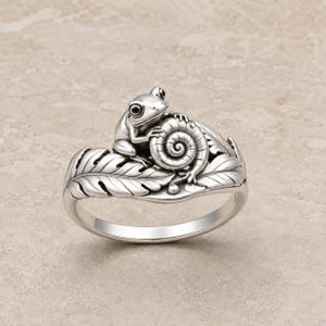 Puede incluir: Un anillo de tono plateado que presenta una rana detallada posada sobre una concha de caracol en espiral, con la banda diseñada como una serie de hojas texturizadas y superpuestas que envuelven el dedo, mostrando metalurgia intrincada.
