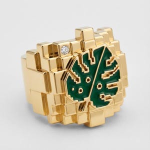 Puede incluir: Un anillo dorado con un diseño pixelado. El anillo presenta un diseño de hoja verde y un pequeño diamante. El anillo tiene una apariencia geométrica y en bloques.