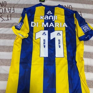 May include: Yellow and blue soccer jersey with the name "DI MARIA" and the number "11" on the back. The jersey has vertical stripes and the text "kanji" on the front.