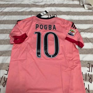 May include: A pink soccer jersey with the name "POGBA" and the number "10" in black. The jersey has black accents on the collar and sleeves, with a team logo on the sleeve. The jersey is made of a lightweight, breathable material.