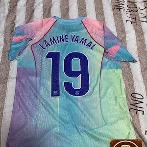 May include: A colorful soccer jersey with the name "LAMINE YAMAL" and the number "19" in navy blue. The jersey has a gradient design with shades of blue, pink, and purple. The jersey is displayed on a striped surface.