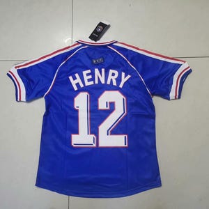 May include: A blue football shirt with the name "HENRY" and the number "12" in white. The shirt has red and white trim on the sleeves and collar. A tag is attached at the top.