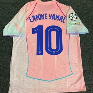 May include: Pink soccer jersey with the name "LAMINE YAMAL" and the number "10" in blue. The jersey has a soccer ball logo and the words "UEFA FOUNDATION for children" on the right sleeve. The sleeves have a pastel color scheme.