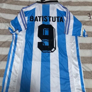 May include: A light blue and white striped soccer jersey with the name "BATISTUTA" and the number "9" on the back. The jersey has a black collar and a tag attached.