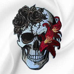 May include: An embroidered patch featuring a light blue skull adorned with dark brown roses on the forehead and a vibrant red lily covering the right eye socket. The design includes black stitching for the teeth, nose, and eye details.