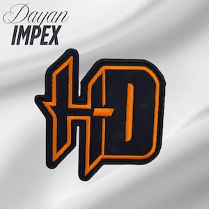 May include: Black and orange embroidered patch featuring the letters "HD" in a stylized design. The patch has a black background with orange outlining and details. The text "Dayan IMPEX" is in the upper left corner.
