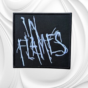 May include: A square-shaped black embroidered patch featuring the text "IN FLAMES" in a stylized, jagged, light blue font. The embroidery has a textured appearance with sharp, branch-like lines extending from the letters, creating a distinct graphic design.