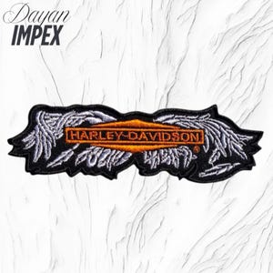 May include: Black embroidered patch featuring the Harley-Davidson logo in orange and white, with a black border and white wing-like designs on each side. The patch has the text "HARLEY-DAVIDSON" in an orange rectangle.
