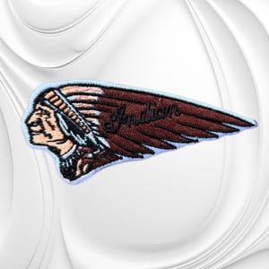 May include: An embroidered patch featuring a profile of a person wearing a feathered headdress in shades of brown, tan, and black. The word Indian is stitched in black cursive script across the dark brown feathers, all outlined in light blue.