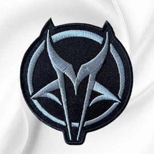 May include: A circular embroidered patch featuring a black background with a stylized, light gray wolf head silhouette integrated into a circular frame. The design uses dense stitching to create sharp, angular lines and a distinct, symmetrical emblem.