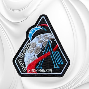 May include: A triangular embroidered patch with a black border featuring a gray moon, a blue stylized letter A, and a red swoosh. The text reads WISEMAN, GLOVER, KOCH, and HANSEN in white lettering against the black background of the patch.