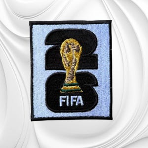 May include: A rectangular embroidered patch featuring a light blue background with a black border. The design displays the black numbers 2026, with a gold and green FIFA World Cup trophy icon centered over the numbers and the white text FIFA below.