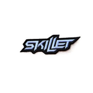 May include: An embroidered patch featuring the word SKILLET in a stylized, angular font. The lettering is light blue with a thick black border, creating a sharp, geometric shape that follows the contours of the text for a bold appearance.