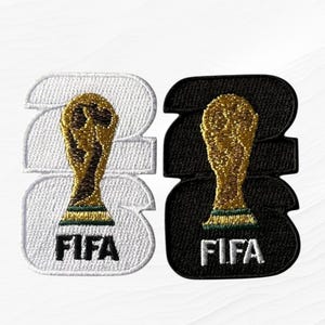 May include: Two embroidered patches featuring the FIFA World Cup trophy. One patch is white with a gold trophy and black text, the other is black with a gold trophy and white text. The patches are in the shape of the number 23.