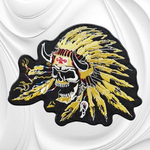 May include: An embroidered patch featuring a skull wearing a large yellow feathered headdress with black horns. The skull is white with black shading, and the headdress includes a central band with a red and white cross symbol and dangling feathers.