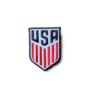 May include: A shield-shaped embroidered patch featuring the letters USA in dark blue at the top, positioned above a series of vertical red stripes on a light blue background, all enclosed within a dark blue border with a pointed bottom edge.