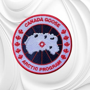 May include: A circular embroidered patch featuring a light blue map of the Arctic on a dark blue background, surrounded by a red border with the text CANADA GOOSE ARCTIC PROGRAM and small red maple leaf icons stitched around the perimeter.