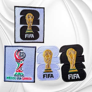 May include: Four embroidered patches featuring FIFA World Cup 2026 branding. One rectangular patch shows a black 26 with a gold trophy and FIFA text. Another shows the 2026 logo with Mexico USA Canada text. Two others feature the trophy on 26.
