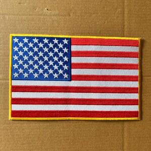 May include: A rectangular embroidered patch featuring the flag of the United States of America. The design includes a blue canton with white stars and alternating red and white horizontal stripes, all bordered by a thin yellow stitched edge.