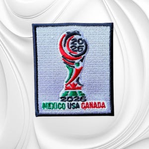 May include: A rectangular embroidered patch featuring a stylized trophy design in red, green, and black on a light gray background. The text 2026 is embroidered at the top and bottom, with the words MEXICO USA CANADA stitched in colored thread.
